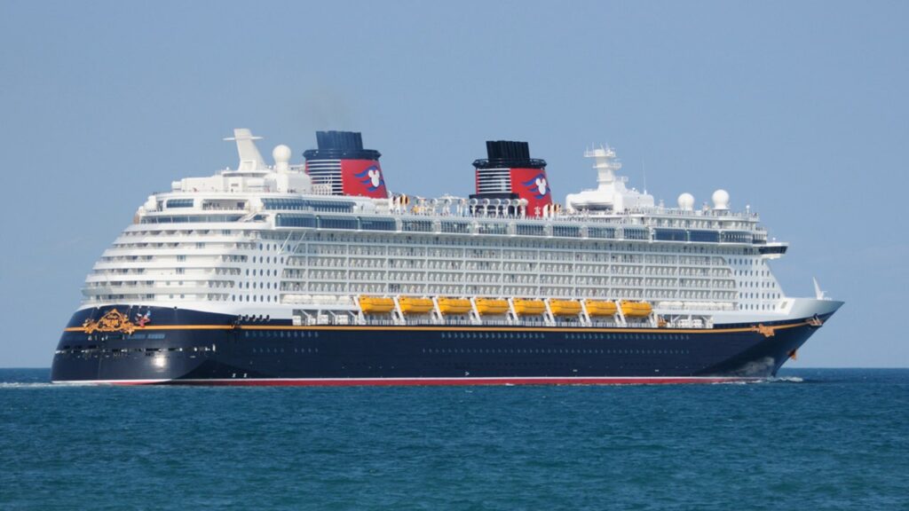 Disney cruise ship with two red and blue funnels sailing on the blue ocean, wake behind it.
