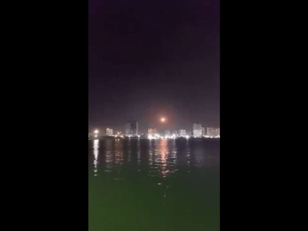 Nighttime city skyline along a body of water with buildings lit and reflecting on the surface.