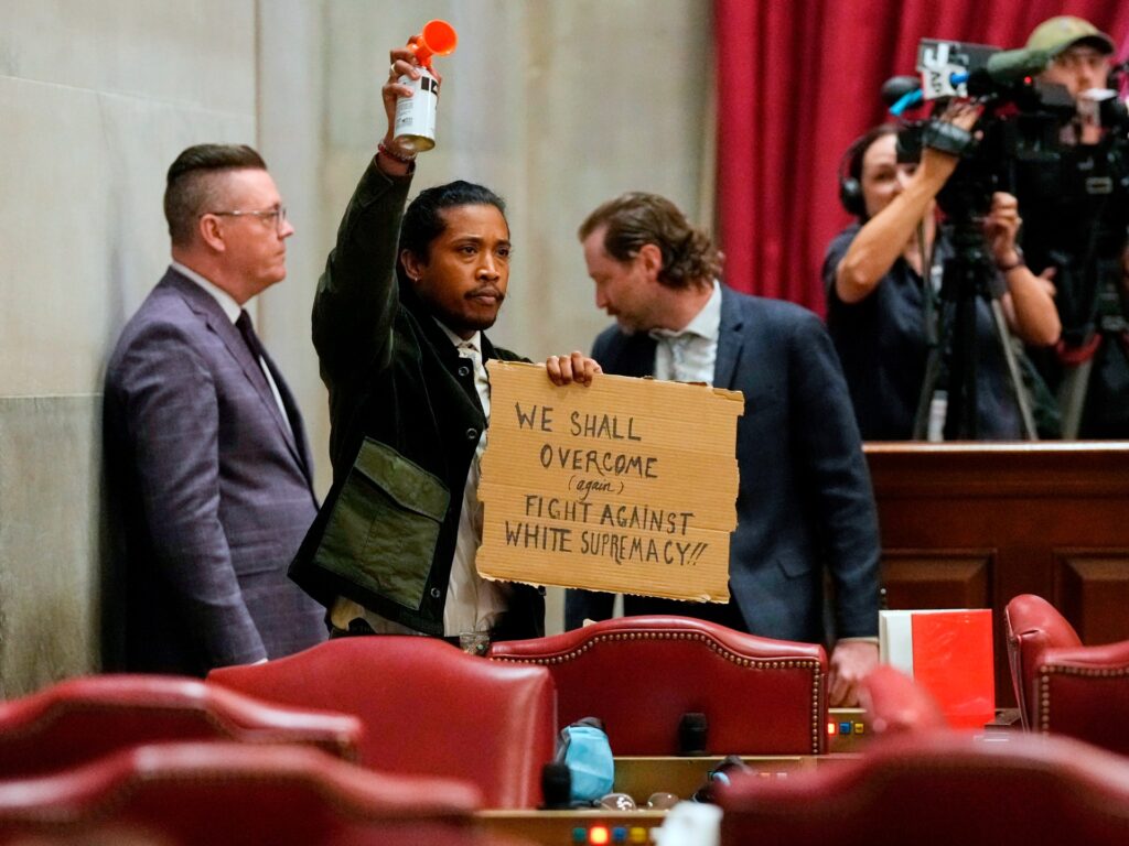 Protester in a dark jacket raises an orange bottle while holding a cardboard sign about fighting white supremacy; a camera crew films nearby in a courtroom setting.
